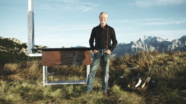 Kurt Gellner steht vor seiner Holzfeuerküche mit Bergen im Hintergrund.